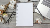 Leerer Schreibblock liegt auf einem Holztisch neben Baumwollknospen, einer Brille und einem Laptop, auf dem ein Bleistift liegt.