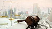 Richterhammer liegt auf der Tastatur eines Laptops. Im Hintergrund ist eine Waage vor der Skyline einer Stadt zusehen.