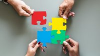 Vier Hände halten vier verschiedenfarbige Puzzelteile aneinander und bilden ein Quadrat