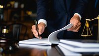 Hand unterschreibt einen Vertrag mit einem Füller. Neben dem Vertrag sind ein Richterhammer und eine Waage zu sehen.